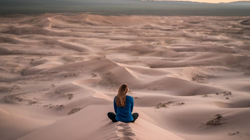 Woman sitting facing sand dunes and horizon. Photo.